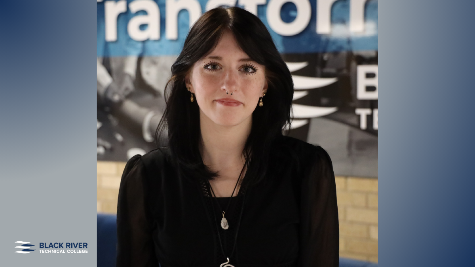 A young woman with long dark hair, wearing a black blouse and layered necklaces, poses in front of a Black River Technical College banner.